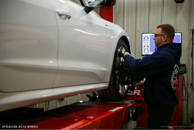 Technician performing a four-wheel alignment on a white sedan, alignment readings shown on the shop monitor behind him.