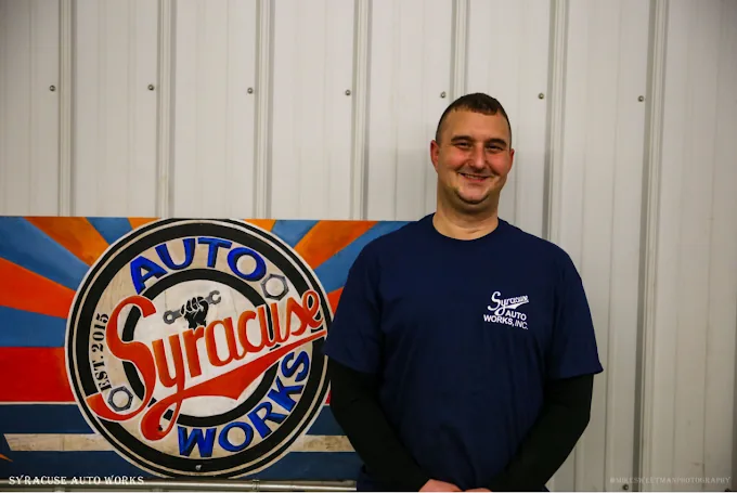 Tim, owner of Syracuse Auto Works, standing in front of the hand-painted shop sign.